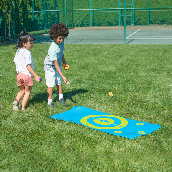 Deux enfants lancent des sacs sur un tapis d'activité, dans un champ d'herbe près d'un court de tennis.