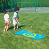 Two kids tossing toss bags at activity mat, in a grass field next to a tennis court
